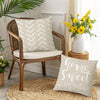A beige woven chair with a white cushion and decorative pillows sits on a round jute mat.