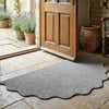 A natural jute non-slip entrance door mat placed on a stone floor near a wooden door and potted plants.