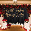 A wedding backdrop featuring a dark blue curtain draped with red floral garlands and gold script reading Will You Marry Me