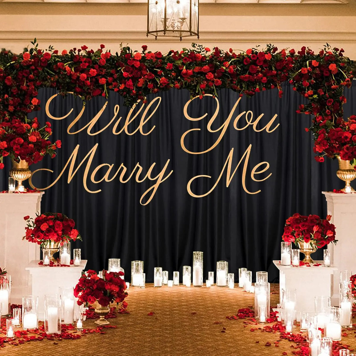 A wedding backdrop featuring a dark blue curtain draped with red floral garlands and gold script reading Will You Marry Me