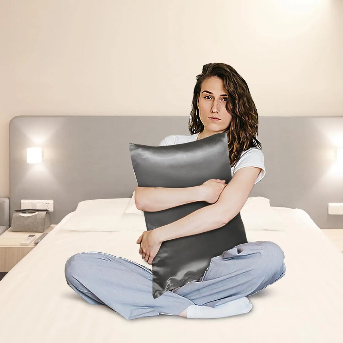 A woman lies on a bed wearing light blue pants and a gray satin pillowcase set.