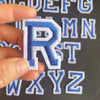 A hand holds a blue embroidered letter R patch against a background of other alphabet patches.