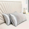 A silver satin pillowcase with an envelope closure resting on a white bed, next to a lamp.