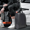 A dark gray portable shoe carrier bag resting on a black mat next to a person's legs and shoes.