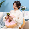 A woman is holding a pink fried doughnut plush pillow while sitting on a couch.