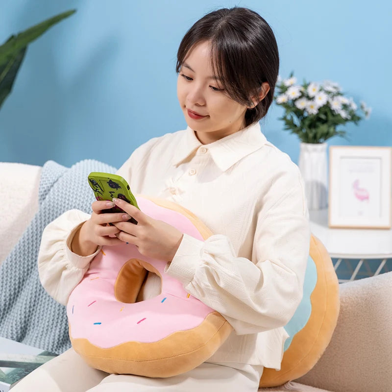 A woman is holding a pink fried doughnut plush pillow while sitting on a couch.