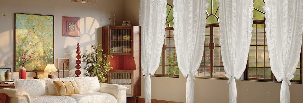 White floral lace curtains hanging in a sunlit room with wooden furniture and large windows.