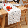 A white Halloween themed table runner featuring ghost patterns and orange pumpkins on a wooden table.