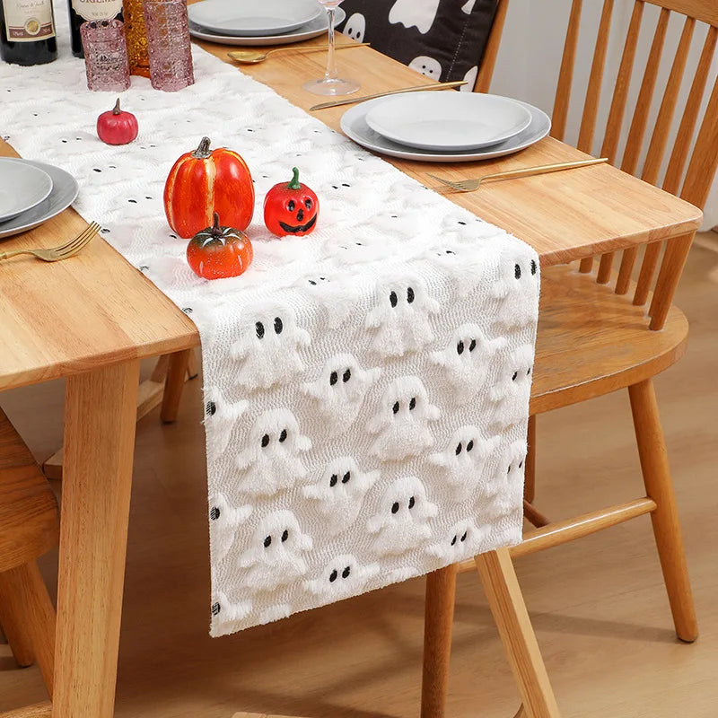A white Halloween themed table runner featuring ghost patterns and orange pumpkins on a wooden table.