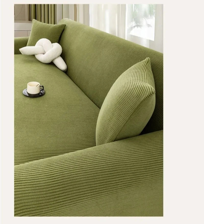 A moss green stretch jacquard sofa slipcover with a cream accent pillow and a cup on the surface.