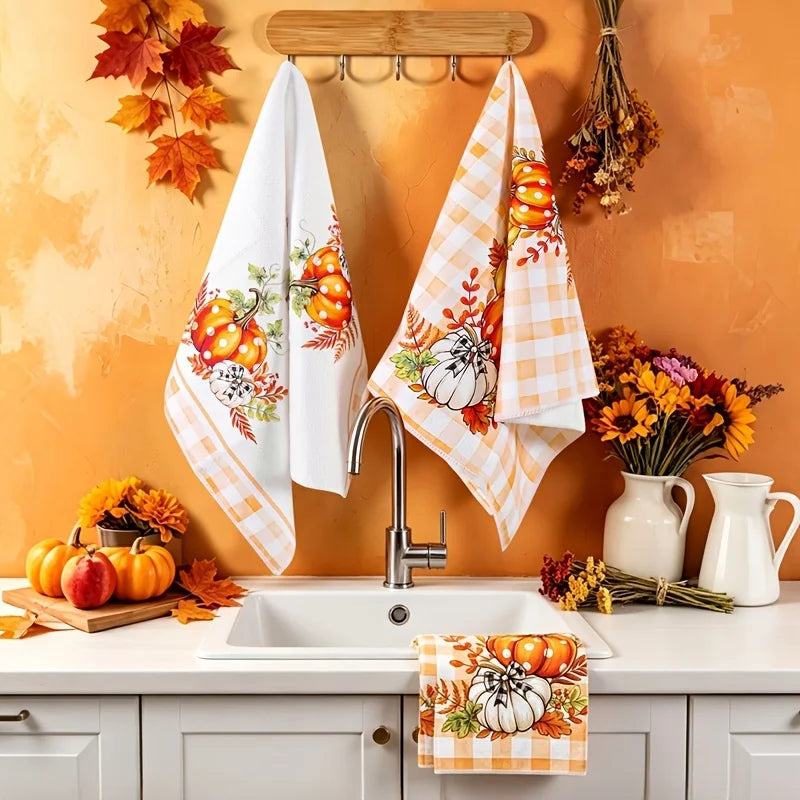 A set of two kitchen towels with a pumpkin and plaid design hanging on a hook against an orange wall.