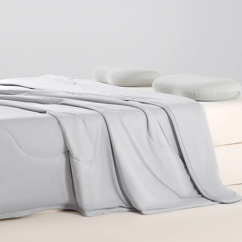 A white ice silk summer cooling blanket is draped over a surface next to a soft white pillow.