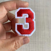 A hand holds a customizable number patch featuring the number three in red and white embroidery.