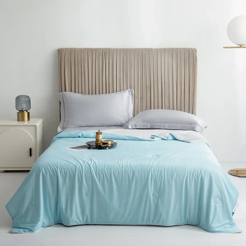 A light blue ice silk cooling comforter is laid on a bed with a neutral upholstered headboard.