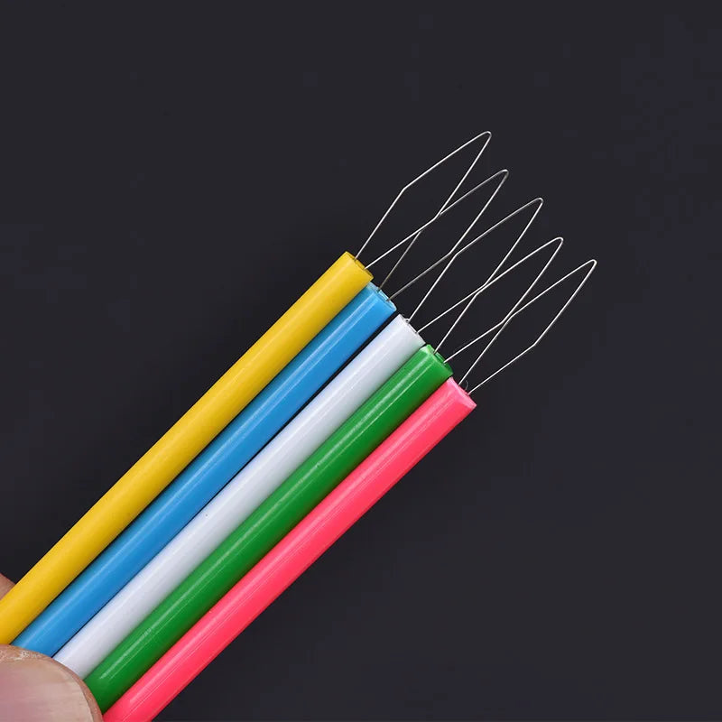 A hand holds a bundle of colorful needle threaders with various colored needles against a dark background.