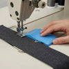 A hand places a blue magnetic seam guide with a stitch gauge onto a dark gray fabric for sewing.