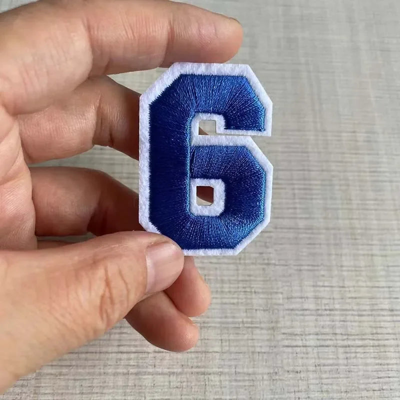 A hand holds a dark blue embroidered number 6 patch with a white border.