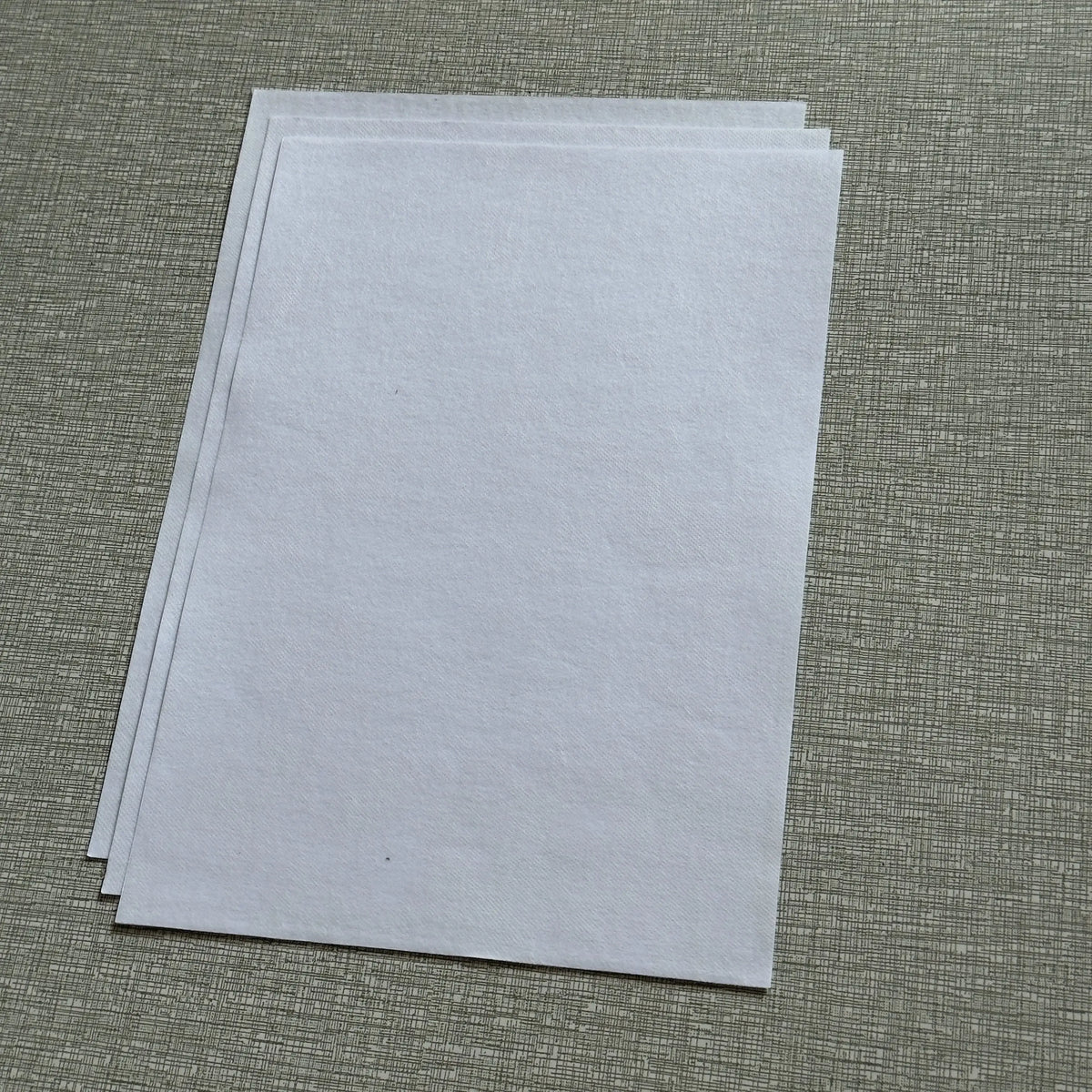 A stack of white water-soluble adhesive embroidery stabilizer sheets lying on a textured green surface.