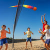 A child is throwing a rainbow comet ball with a soft tail high into the air against a clear blue sky on a sandy beach.