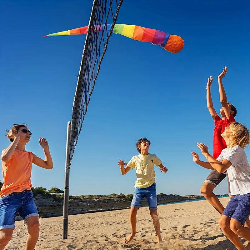 A child is throwing a rainbow comet ball with a soft tail high into the air against a clear blue sky on a sandy beach.
