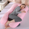 A woman lying on a pink U-shaped maternity body pillow in a light pink bedroom setting.
