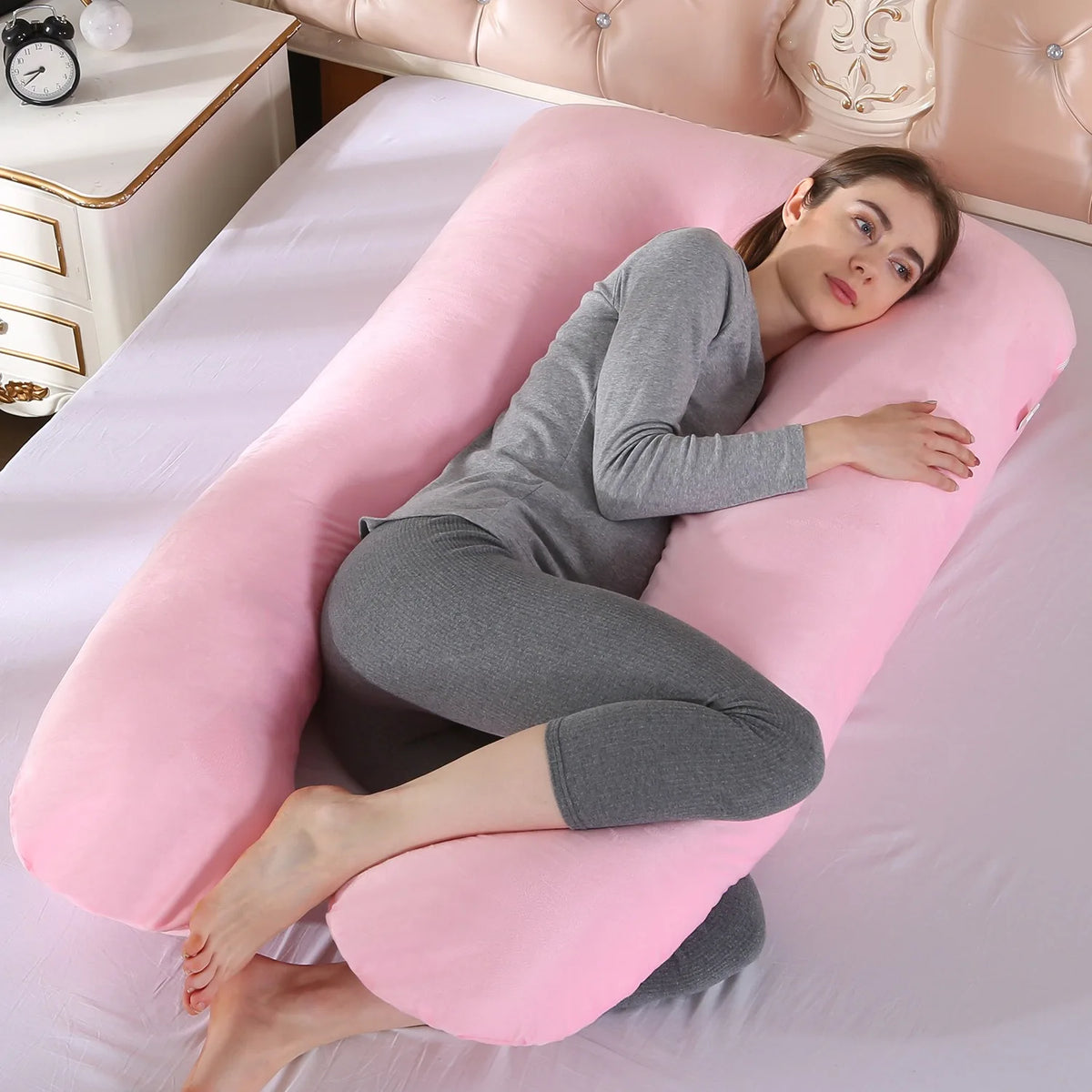 A woman lying on a pink U-shaped maternity body pillow in a light pink bedroom setting.
