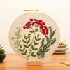 A circular embroidery hoop featuring floral cross stitch designs in shades of green and red on a beige fabric.