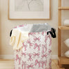 A pink and white canvas laundry basket decorated with a pattern of pink bows, shown on a wooden floor.