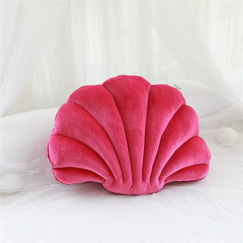 A pink velvet stuffed sofa cushion shaped like a decorative seashell resting on white fabric.