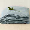 Light blue pure linen flat sheet folded and draped on a light surface with a green leaf.