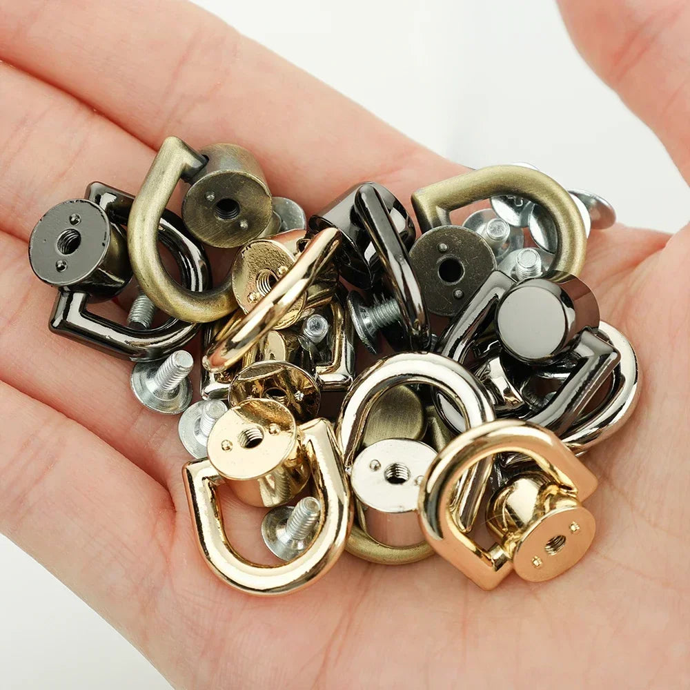 A collection of metal D-shaped ring bag handles and screws in various gold and silver tones held in a hand.