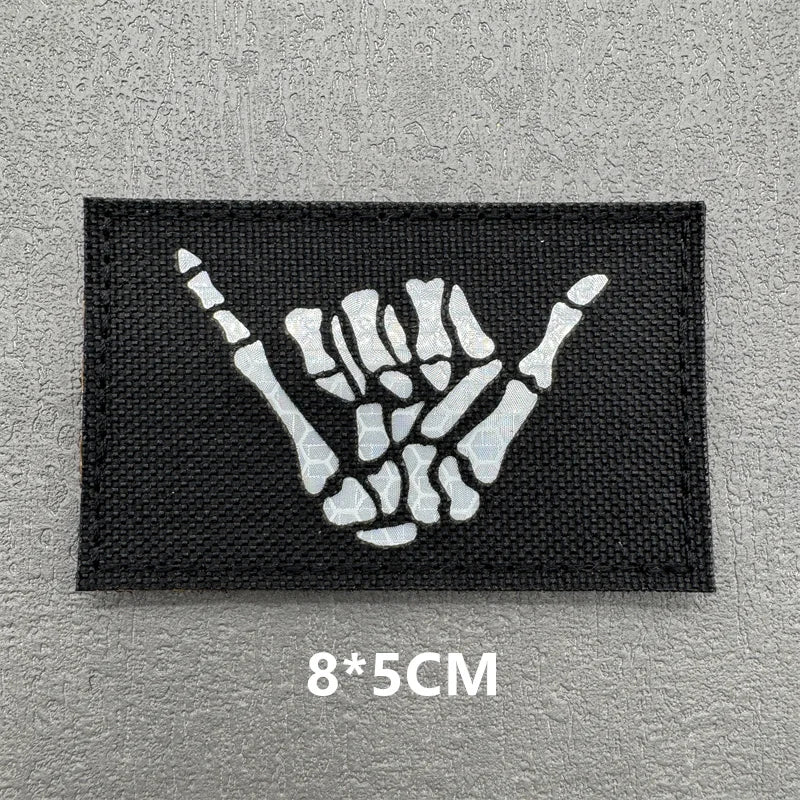 A black embroidered patch featuring a white hand gesture symbol on a gray background.