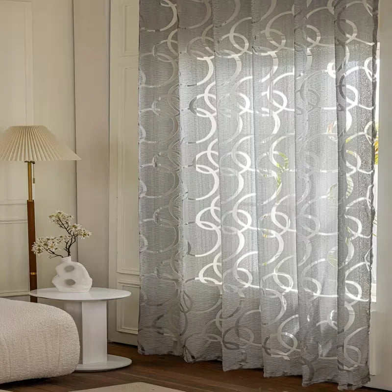 A floor-to-ceiling sheer curtain with a gray geometric embroidered pattern hangs in a bright room.