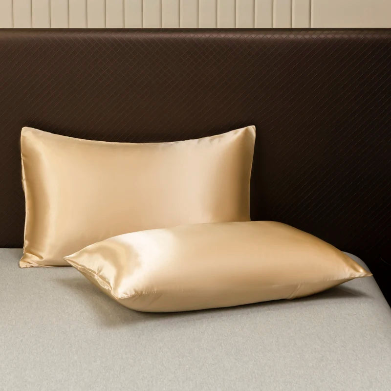 A pair of smooth satin pillows resting on a dark brown headboard and light gray bedding.