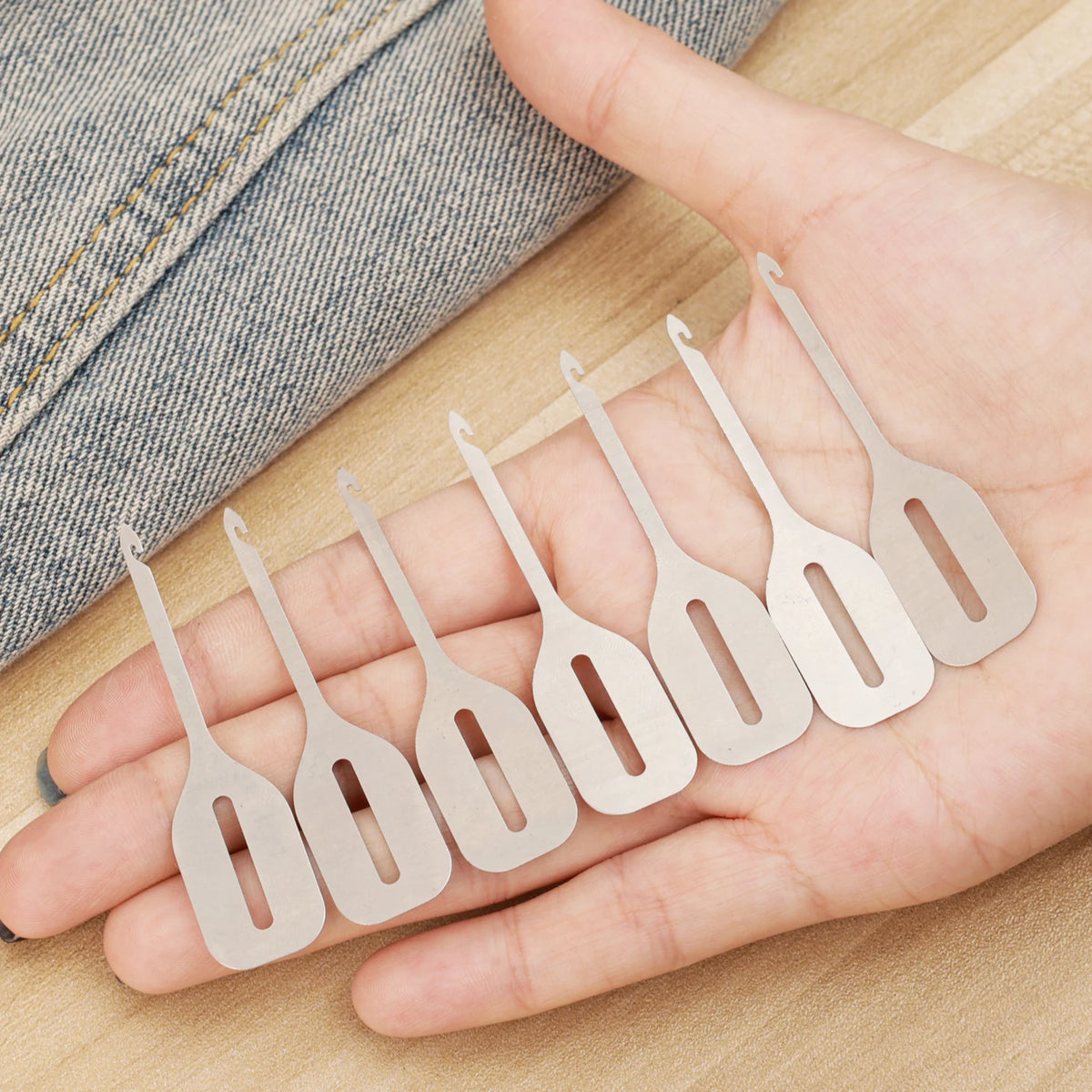 A hand holds a set of stainless steel needle threaders for hand sewing and embroidery on a wooden surface.