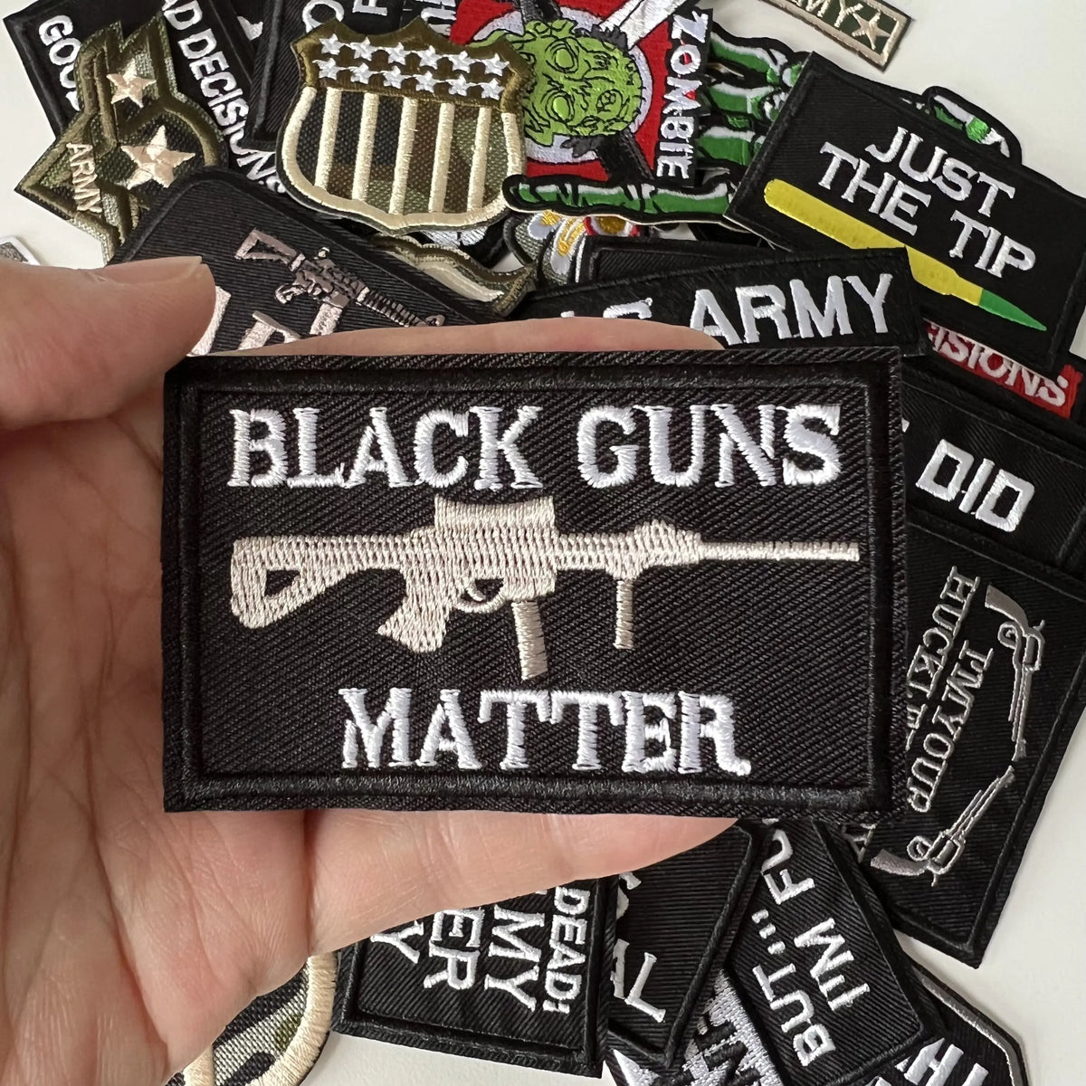A hand holds a black iron-on patch with the text BLACK GUNS MATTER on a dark fabric background.