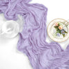 A flowing lilac cheesecloth table runner draped elegantly with a white glass and decorative floral accents.