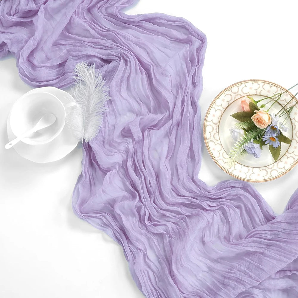 A flowing lilac cheesecloth table runner draped elegantly with a white glass and decorative floral accents.