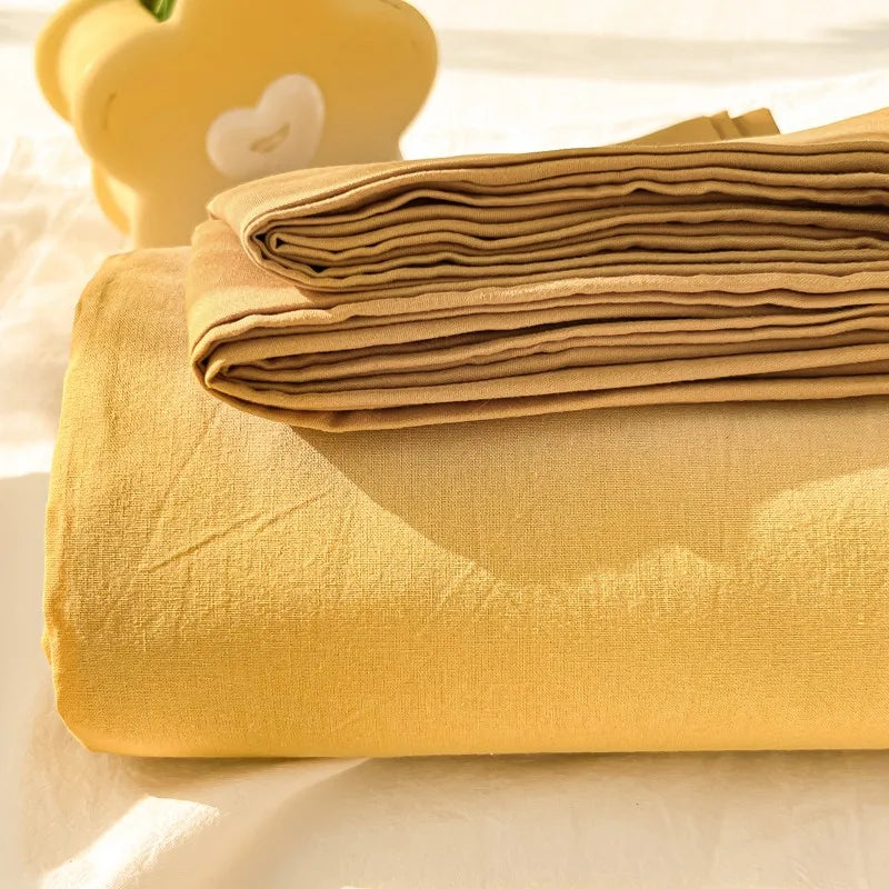 A stack of mustard yellow organic cotton bedding, including a folded flat sheet and pillowcases, next to a light yellow decor