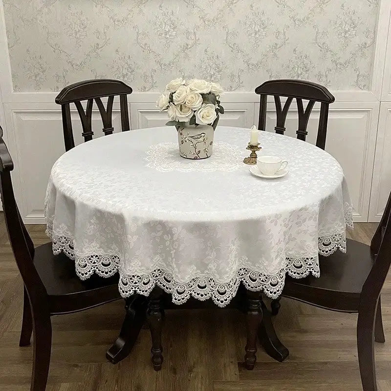 A round white embroidered lace tablecloth set for a meal with a vase of white roses and a teacup.