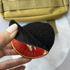 A hand holds a black embroidered patch with red lettering and a gold hook and loop backing.