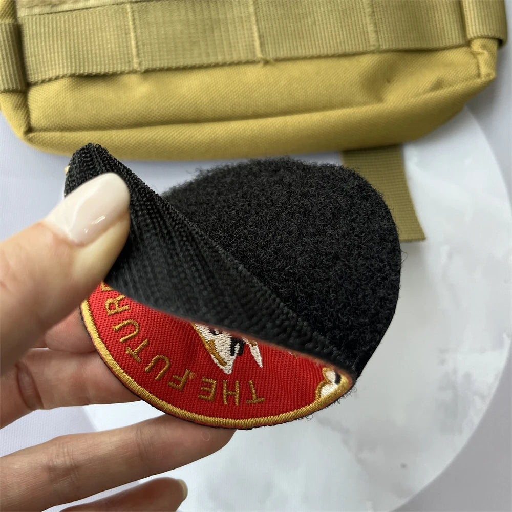 A hand holds a black embroidered patch with red lettering and a gold hook and loop backing.