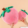 A pink plush strawberry throw pillow with green leaves and a brown stem being held by hands.