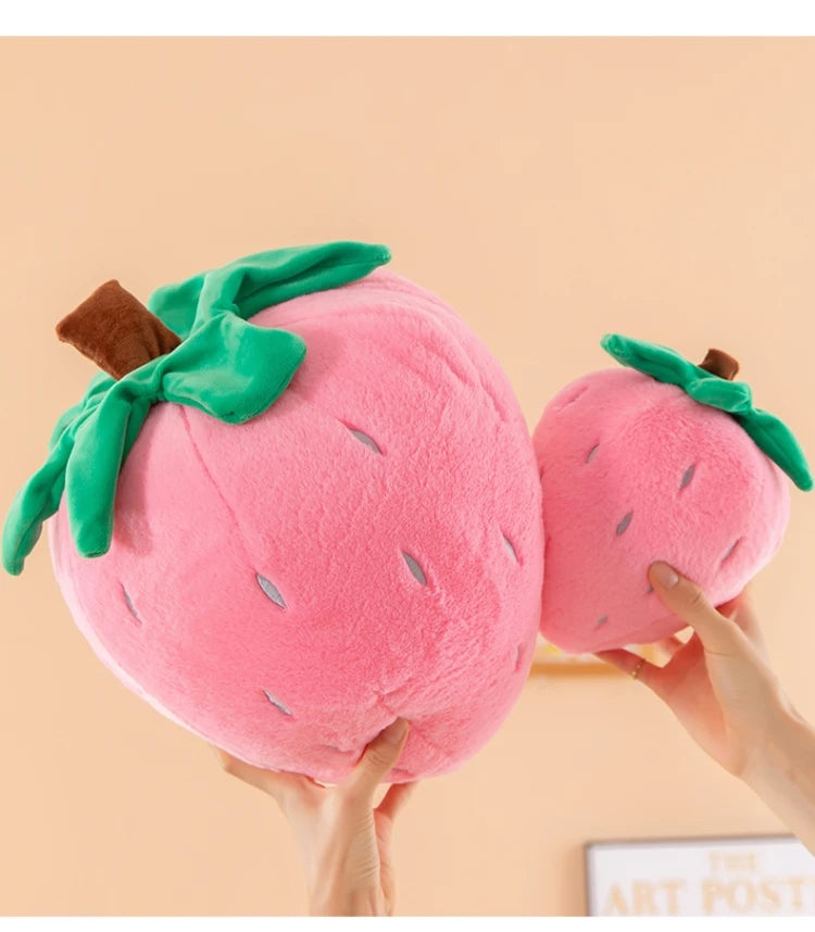A pink plush strawberry throw pillow with green leaves and a brown stem being held by hands.