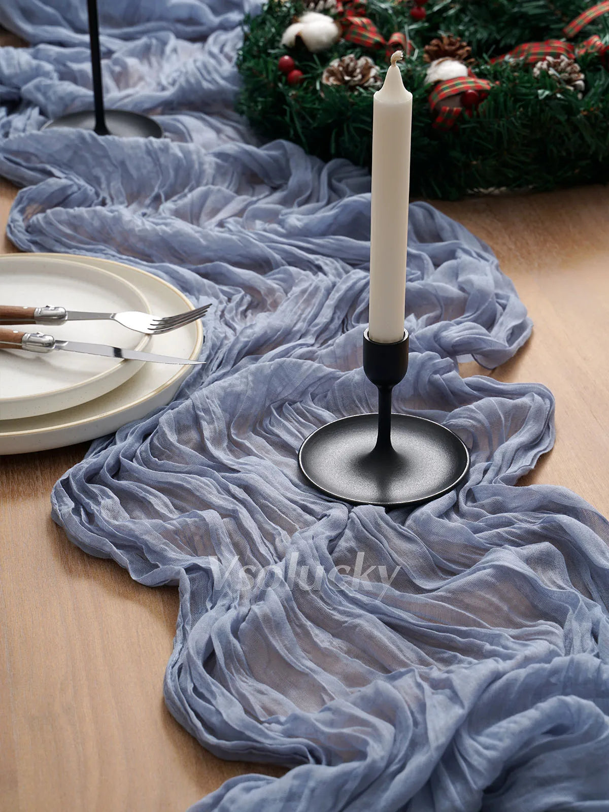 A grey crinkled cheesecloth table runner draped over a wooden surface with a white candle and greenery.