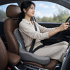 A woman is sitting in a car, driving, wearing a beige shirt and pants, with an ergonomic memory foam car seat cushion visible
