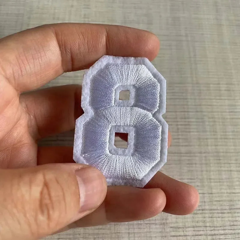 A hand holds a white embroidered number patch shaped like the numeral 8.