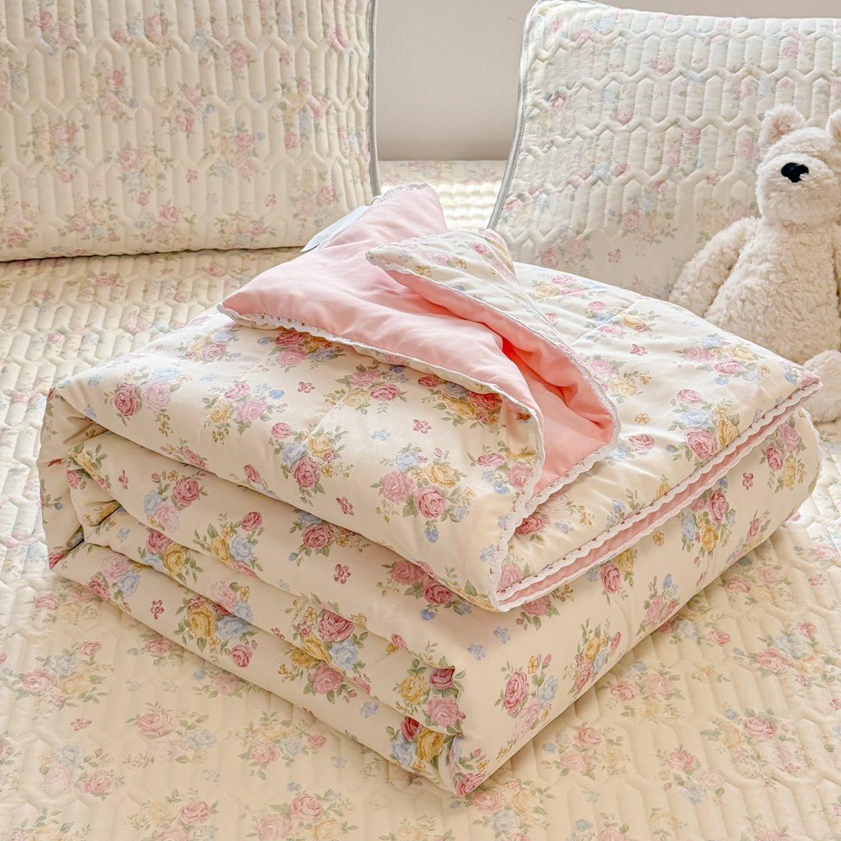 A stack of pink and cream quilted blankets resting on a light patterned fabric with a white stuffed animal in the background.