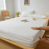 A white fitted sheet is laid out on a wooden bed frame with a hand resting on the fabric.