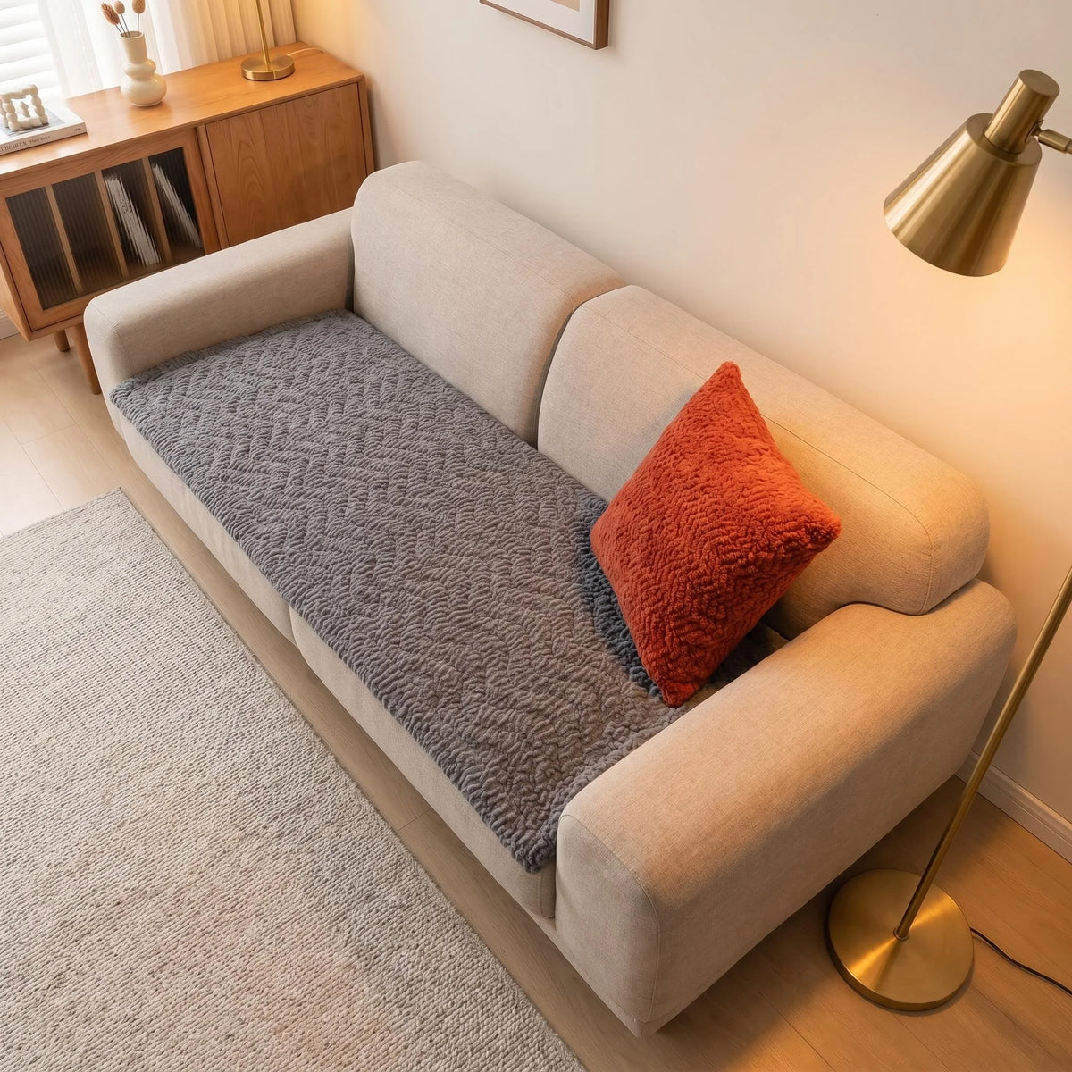 A light beige sofa with a gray flannel pad cover and an orange accent pillow sits on a light rug.